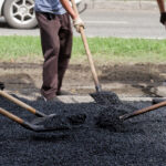 Close view of workers raking and shoveling new blacktop by the curb; team effort in street repair and urban improvement.