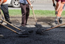 Close view of workers raking and shoveling new blacktop by the curb; team effort in street repair and urban improvement.