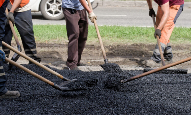 Close view of workers raking and shoveling new blacktop by the curb; team effort in street repair and urban improvement.
