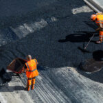 Construction workers wearing orange safety gear are paving a road with fresh asphalt.