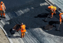 Construction workers wearing orange safety gear are paving a road with fresh asphalt.