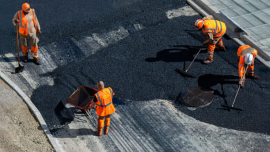 Construction workers wearing orange safety gear are paving a road with fresh asphalt.