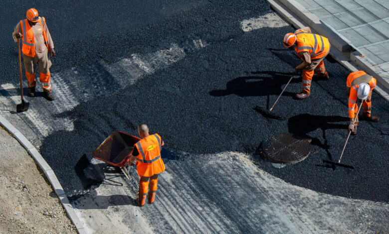 Construction workers wearing orange safety gear are paving a road with fresh asphalt.